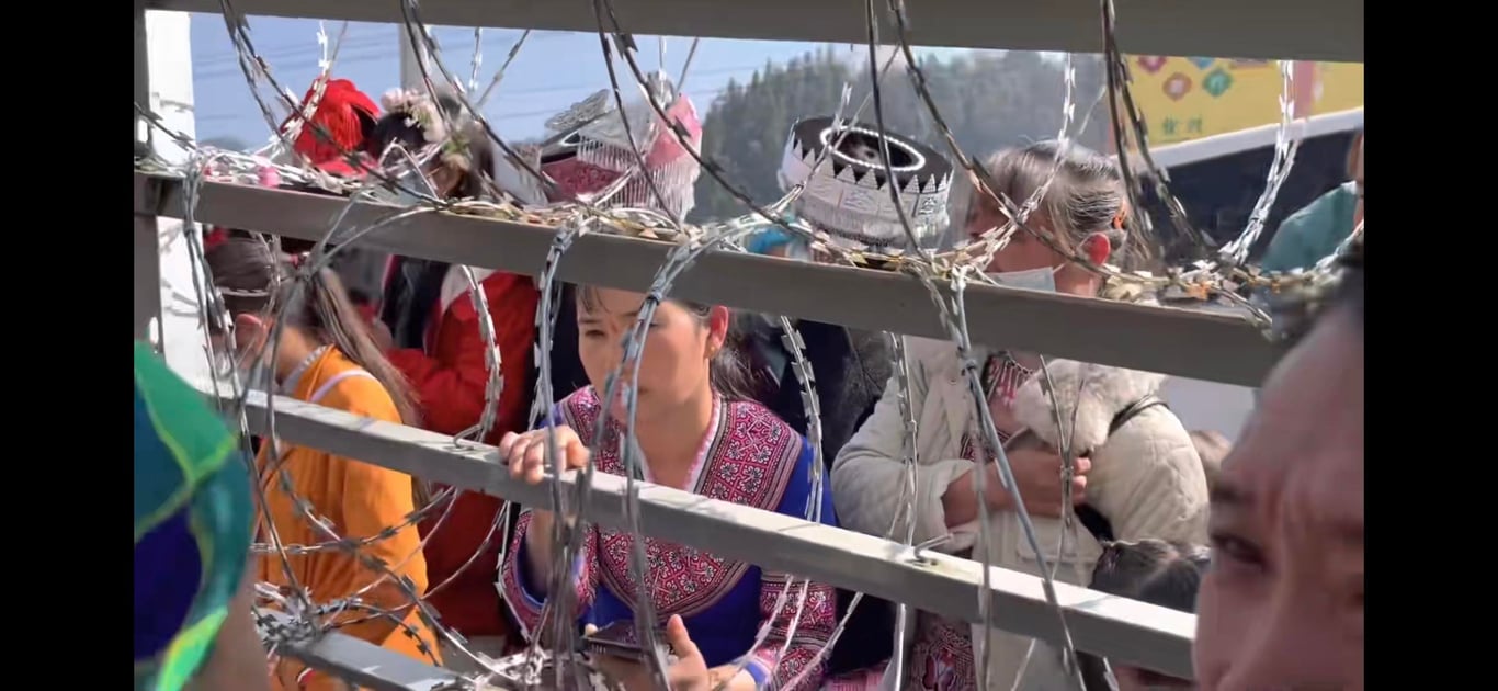 A group of women wearing colorful clothing and headdresses, seen through metal bars and barbed wire.