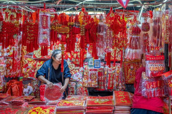 The proprietor of a small stall selling colorful red Chinese Lunar New Year decorations (many adorned with dragons, in celebration of The Year of the Dragon 2024) gives a big smile to a person in a red jacket standing on the other side of the stall.